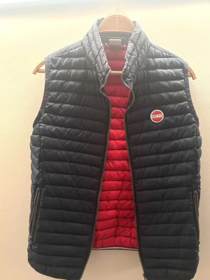 Svart dunväst från Colmar - Säljer en svart quiltad dunväst från Colmar med röd insida och hög krage. Västen har två fickor med dragkedja och ett Colmar-märke på bröstet. Perfekt att slänga över en hoodie eller tröja för extra värme. Pris kan diskuteras 