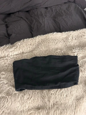 Svart bandeau tubtopp - Enkel och stilren svart bandeau tubtopp i mjukt stretchigt material. Perfekt att styla med höga jeans eller kjol för en clean look. Axelbandslös design som sitter bekvämt på plats.