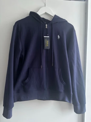 Hoodie Polo Ralph Lauren med prislappar 🧥 - Det här är en marinblå hoodie från Polo Ralph Lauren, med den ikoniska logotypen på bröstet. Plagget är helt nytt med prislappar kvar.  ⭐️ Skick: 10/10 🎲 Storlek: M 🎨 Färg: Marinblå 📦 Snabb och varsam frakt
