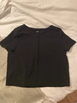 Svart blus med knappar från Gina Tricot - Säljer en stilren svart blus från Gina Tricot i storlek 36. Blusen har korta ärmar, rund halsringning och dekorativa knappar framtill. Materialet känns mjukt och har en lätt struktur, perfekt för att skapa en clean look.