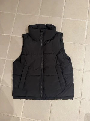 Svart dunväst med hög krage - Säljer en svart dunväst med hög krage och dragkedja framtill. Västen har två snedställda fickor och quiltad design som ger en schysst look. Perfekt att slänga över en hoodie eller tröja för extra värme.