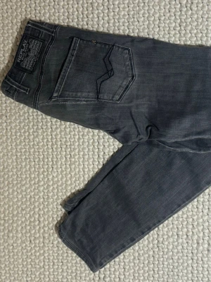 Grå Replay anbass jeans - Säljer ett par grå jeans från Replay med klassisk femficksdesign. Jeansen har tydliga sömmar och en snygg tvättad look. Märkespatch bak i midjan och diskreta detaljer på bakfickorna. Perfekta för dig som gillar stilrena och bekväma jeans. Har lite slitage och skulle nog behöva sy lite, därav priset !