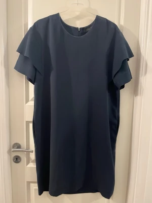 Mörkblå klänning med volangärm från COS - Säljer en stilren mörkblå klänning från COS i storlek 44. Klänningen har rund halsringning, korta volangärmar och rak passform. Materialet känns mjukt och har ett snyggt fall. Perfekt för dig som gillar minimalistisk design.
