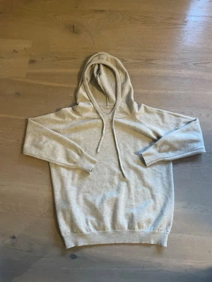 Beige merinoull hoodie  - Säljer en beige hoodie med huva och snörning. Tröjan har långa ärmar, ribbade muddar och en avslappnad passform. Perfekt för dig som gillar en enkel och stilren look. Materialet känns mjukt och skönt mot huden.