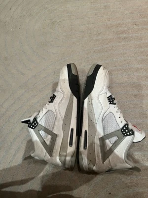 Nike Air Jordan 4  - Nike Air Jordan 4 sneakers i vit och grå färg med svarta detaljer. Skorna har snörning, meshpaneler på sidorna och en ikonisk Jumpman-logga på hälen och tungan. Sulan är platt med synlig luftdämpning och grå stänk. Materialet är skinn och mesh