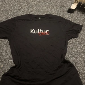 Svart Kultur Crew t-shirt - Svart t-shirt,'Kultur Crew' i vitt och rött på bröstet. Modellen är klassisk med rund hals och korta ärmar. Materialet är mjuk bomull och t-shirten är climate neutral. Perfekt för dig som vill ha en chill och enkel stil. Även väldigt snyggt o ha rött hårband till tröjan 