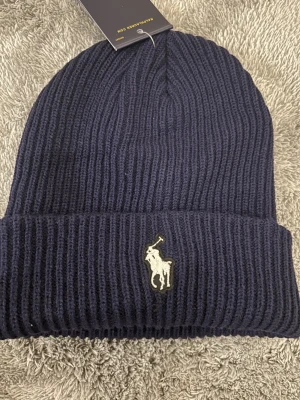 Mörkblå stickad mössa från Polo Ralph Lauren - Snygg mörkblå stickad mössa från Polo Ralph Lauren med klassisk ribbad design och broderad logga framtill. Perfekt accessoar för kyliga dagar och enkel att matcha med din jacka. Materialet är mjukt och värmande, och mössan har en bred uppvikt kant.
