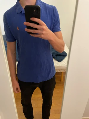 Piké Ralph Lauren - Säljer en klassisk blå pikétröja från Polo Ralph Lauren med orange broderad logga på bröstet. Storleken är S.  Tröjan  har korta ärmar, krage och två knappar vid halsen. Perfekt för dig som gillar stilren och sportig look. Pris går såklart och diskuteras 👍🤗