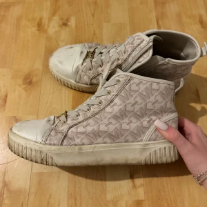 Beige MK sneakers med monogram - Snygga höga sneakers från Michael Kors i beige med MK-monogram över hela skon. Skorna har vita detaljer, guldfärgade logodetaljer på snörningen och dragkedja på sidan. Tjock vit sula och rund tå. Perfekta för dig som gillar streetstyle och vill sticka ut.
