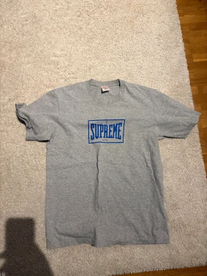 Grå Supreme t-shirt med blå logga - Supreme t-shirt i grått med klassisk blå Supreme NYC-logga på bröstet. T-shirten är i bomull och har en normal passform med korta ärmar. Perfekt för dig som gillar streetwear och vill sticka ut med en ikonisk look.