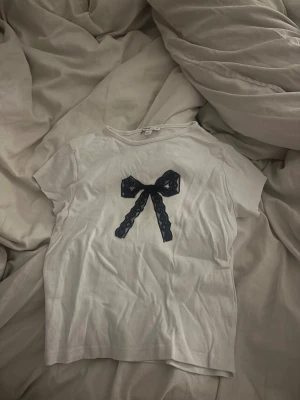 Vit t-shirt med svart rosett Subdued - Vit t-shirt från Subdued i bomull med korta ärmar och en svart spetsrosett framtill. Enkel och söt modell med normal passform, perfekt för en trendig och avslappnad look.