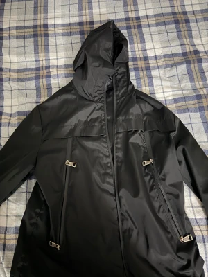 Svart Moncler dunjacka med huva - Svart dunjacka från Moncler med huva och coola dragkedjor framtill. Insidan har Moncler-logga och detaljer i rött samt serietidningsmönster. Jackan har en sleek look och är tillverkad i blankt syntetmaterial med dunfyllning. Perfekt för dig som vill ha en stilren jacka med edge.