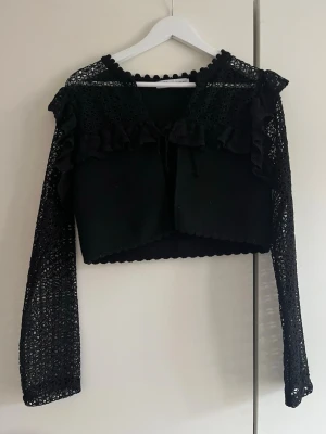 Svart spetskofta från Zara - Säljer en svart kofta från Zara med långa spetsärmar och volangdetaljer över axlarna. Koftan är croppad och har en söt knytning framtill samt dekorativ kant runt halsen. Perfekt för dig som vill ha något unikt och trendigt i garderoben.