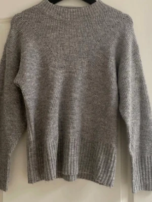 Grå stickad tröja med ribbad struktur - Säljer en grå stickad tröja med ribbad struktur och rund hals. Tröjan har långärmade och är perfekt för kyliga dagar. Enkel och stilren design som passar till jeans eller kjol. Mjuk och skön känsla tack vare det stickade materialet.
