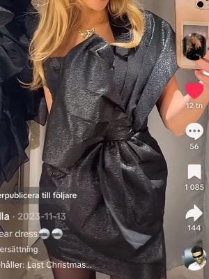 SÖKER! - Säljer en svart glittrig kortklänning med cool omlott-design och stor rosett framtill. Klänningen har en unik struktur och är axelbandslös, perfekt för att sticka ut på festen. Materialet har en skimrande finish som fångar ljuset snyggt.