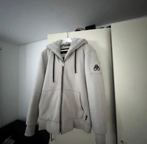 Grå hoodiejacka från Moose Knuckles - Säljer en beige hoodiejacka från Moose Knuckles med dragkedja och svarta snören i huvan. Jackan har två stora fickor framtill och ett snyggt logomärke på vänster ärm. Perfekt för dig som gillar streetwear och vill ha något stilrent och bekvämt. INGET BYTE!