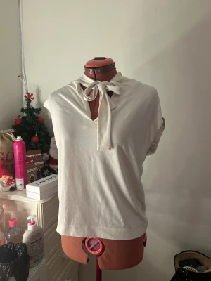 Vit blus med knyt i halsen - Säljer en stilren vit blus med korta ärmar och snygg knytning i halsen. Blusen har en mjuk, slät yta och v-ringad öppning framtill. Perfekt för dig som gillar enkla och eleganta detaljer. Passar till många olika looks och är superenkel att matcha.