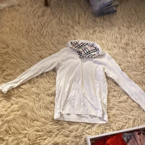 Vit henley från Burberry London - Säljer en vit henley från Burberry London i storlek M. Skjortan har lång ärm och klassisk rutmönstrad insida på kragen i svart, rött och grått. Tillverkad i mjuk bomull och har knappar vid halsen. Perfekt för dig som gillar stilrena och ikoniska detaljer.