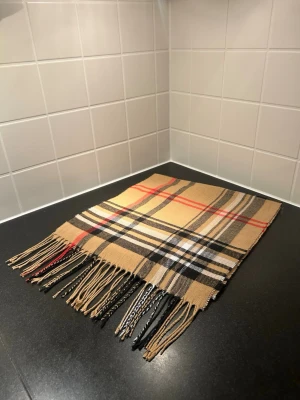 Beige rutig halsduk med fransar - Säljer en beige halsduk med klassiskt rutigt mönster i svart, vitt och rött. Halsduken har snygga fransar i ändarna och är tillverkad i ett mjukt material som känns skönt mot huden. Perfekt att slänga över axlarna för en cool look.