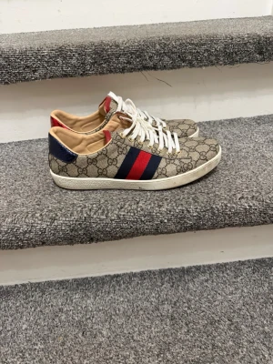 Gucci sneakers med GG-mönster - Säljer ett par snygga Gucci sneakers med klassiskt GG-mönster i beige och brunt. Skorna har vita snören, röd och marinblå rand på sidan samt metallic-detaljer i rött och blått på hälen. Platt vit sula med Gucci-logga undertill. Perfekt för dig som vill sticka ut.