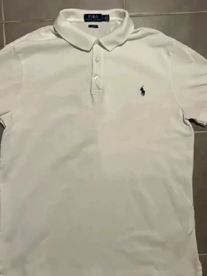 Vit piké från Polo Ralph Lauren - Klassisk vit pikétröja från Polo Ralph Lauren med korta ärmar och krage. Tröjan har tre knappar framtill och den ikoniska broderade loggan i svart på bröstet. Materialet är mjuk bomull som andas och passar perfekt till sommaren.