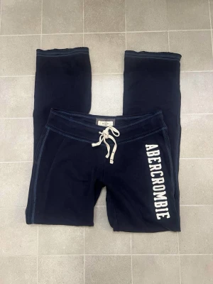 Abercrombie mjukisbyxor mörkblå - Snygga mörkblå/svarta mjukisbyxor från Abercrombie & Fitch med vit snörning i midjan och stort vitt tryck längs benet. Storlek M. Lite urtvättade/noppriga. Vet ej om det är herr/dam. Mått kan skickas.