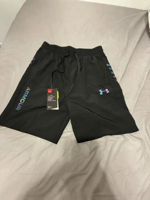 Svarta träningsshorts från Under Armour - Svarta shorts från Under Armour med färgglada logodetaljer på sidan och framtill. De har elastisk midja med snörning för skön passform och är gjorda i lätt polyester som andas. Perfekta för gymmet eller löprundan, riktigt snygga och sportiga. Priset är för hela setet!