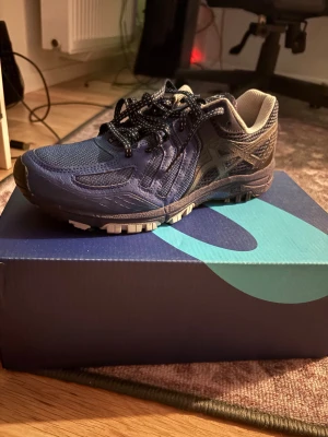 Asics Skor - Säljer ett par blå oanvända Asics sneakers med svart och vit mönstrad snörning. Skorna har meshpaneler för bra ventilation och detaljer i syntetmaterial. Yttersulan är grov för extra grepp och insidan är ljusgrå. Perfekt för dig som gillar sportig stil.