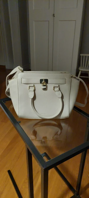 Beige axelremsväska  - Beige axelremsväska med justerbar rem. Syntetläder och gulddetaljer. Två mindre sidofack och en ficka med dragkedja på insidan. Ett ytterfack med dragkedja. Ca. 35 cm bred och 25 cm hög.