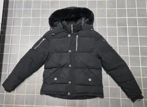 Svart pufferjacka med päls från Moose Knuckles - Snygg svart pufferjacka, Original Shearling Trim 3Q, från Moose Knuckles med fluffig päls på huvan och silvriga detaljer. Jackan har flera dragkedjor, knappar och en ikonisk logga på ärmen. Perfekt för kalla dagar med sin tjocka och quiltade design.