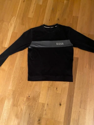 Svart långärmad tröja från BOSS - Svart långärmad tröja från BOSS med en bred grå rand över bröstet och tydlig logotyp. Tröjan har rund hals och är tillverkad i mjuk bomull, perfekt för chill dagar.  Priset är inte fast och kan diskuteras!