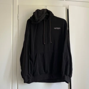 Svart NASA hoodie med tryck - Svart hoodie med stort grafiskt print på ryggen med rymdtema, astronaut-nalle och text. Hoodien har huva med snörning, känguruficka och långa ärmar. Lite streewear vibes