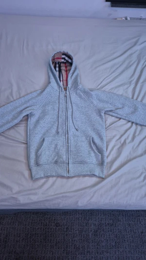 Grå Burberry hoodie med dragkedja - Säljer en grå hoodie från Burberry i storlek S. Tröjan har klassisk dragkedja, snöre vid huvan och Burberrys ikoniska rutiga mönster på insidan av huvan. Mjukt bomullsmaterial och två fickor framtill. Perfekt för chill dagar och snyggt och stiligt