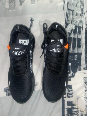 Nike Air 270 x Off-White svarta sneakers - Svarta Nike Air 270 sneakers i samarbete med Off-White. Skorna har vit text, orange detalj och tydliga Off-White-prints. Snygg mesh-ovandel, snörning och platt sula med synlig Air-enhet. Perfekt för dig som gillar streetwear och unika detaljer.