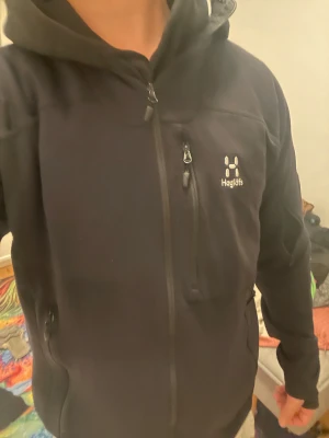 Svart fleecejacka från Haglöfs M - Svart fleecejacka från Haglöfs i storlek M. Jackan har huva, flera dragkedjeförsedda fickor och ett diskret Haglöfs-logo på bröstet. Materialet är mjukt och värmande fleece med en lätt ribbad struktur. Perfekt för utomhusaktiviteter och chill.