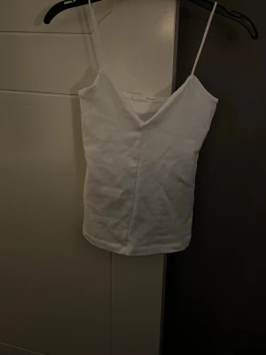 Vit linnetopp med smala axelband - En stilren vit linnetopp med smala axelband och v-ringning. Toppen är enkel och passar perfekt till sommaren, med en rak passform och mjukt material. Perfekt att matcha med jeans eller kjol för en fräsch look.