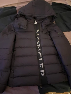 Moncler jacka black label - Säljer en svart dunjacka från Moncler med stor logga längs dragkedjan och klassisk badge på ärmen. Jackan har huva med knappar, med tags o allt kvar. Insidan är glansig och jackan är perfekt för kalla dagar.