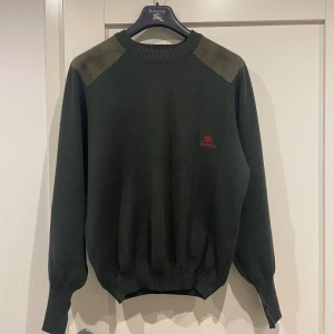 Mörkgrön stickad tröja Burberry - Snygg mörkgrön stickad tröja från Burberrys med broderad röd logga på bröstet. Tröjan har mockadetaljer på axlar och armbågar, ribbad hals och muddar. Tillverkad i 100% ren ull, perfekt för kyliga dagar. Klassisk och stilren design.