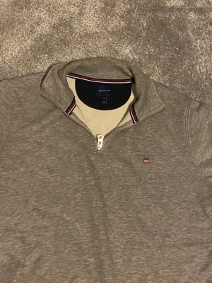 Grå half zip tröja från GANT - Snygg grå långärmad tröja från GANT med half zip och ståkrage. Tröjan har ett litet broderat GANT-emblem på bröstet och diskreta ränder på insidan av kragen. Perfekt för dig som gillar stilren och klassisk design.