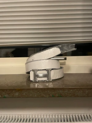 Vit skinnbälte med H-spänne - Snyggt vitt bälte i skinn med ett stort H-format metallspänne. Bältet har en stilren och modern look med diskreta svarta sömmar längs kanterna. Perfekt för att lyfta din outfit med en clean och exklusiv vibe. Kan bli vit svart