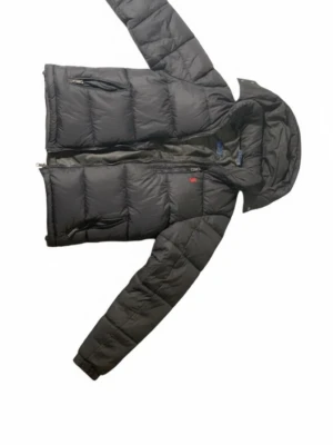 Svart dunjacka med huva - Säljer en svart dunjacka från Peak Performance med huva och flera praktiska fickor med dragkedja. Jackan har quiltad design och långärmade ärmar, perfekt för kalla dagar. Snygg och funktionell för dig som vill ha värme och stil.