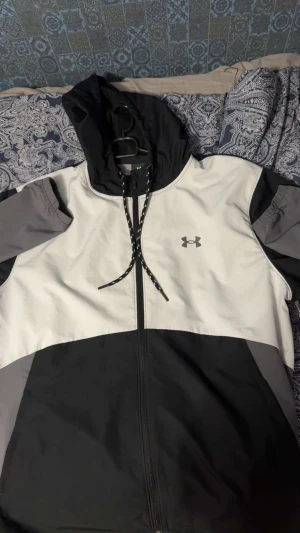 Under Armour vindjacka XL svart/vit - Snygg vindjacka från Under Armour i färgerna svart, vit och grå. Jackan har huva med snörning, hel dragkedja framtill och meshfoder på insidan. Perfekt för dig som gillar sportig stil och vill ha något lätt och funktionellt.