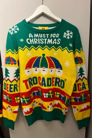 Jultröja Trocadero S färgglad - Färgglad jultröja från Trocadero i storlek S med grönt, gult, rött och blått. Stickad med motiv av tomtar, snögubbar, julklappar och snöflingor samt texten 'A must for Christmas'. Perfekt för att sticka ut på julfirandet! Har en liten defekt men inget som syns väl.