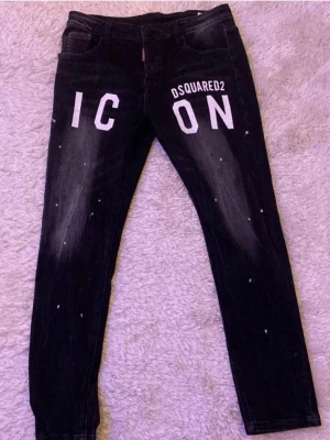 Svarta Dsquared2 ICON jeans - Säljer ett par svarta jeans från Dsquared2 med stor vit ICON-print framtill och coola slitningar samt färgstänk. Jeansen har klassisk femficksdesign, silverfärgad ringdetalj bak och patch med Dsquared2-logga. Passformen är slim och materialet är jeans.