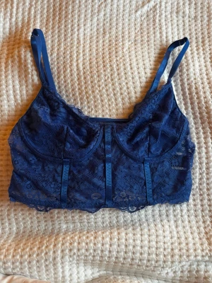 Blå spetskorsett med smala axelband - Säljer en marinblå korsettopp i spets med smala justerbara axelband. Toppen har vackra spetsdetaljer och dekorativa sömmar framtill. Perfekt att styla till jeans eller kjol för en trendig look.