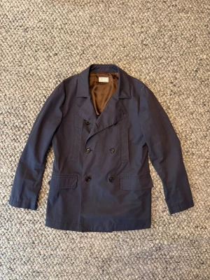 Brunello Cucinelli jacka  - Säljer en stilren mörkblå jacka från Brunello Cucinelli, tillverkad i Italien. Jackan har dubbelknäppning, klassisk krage och två stora fickor framtill. Insidan är fodrad med brunt, glansigt tyg och har snygga detaljer vid innerfickorna.