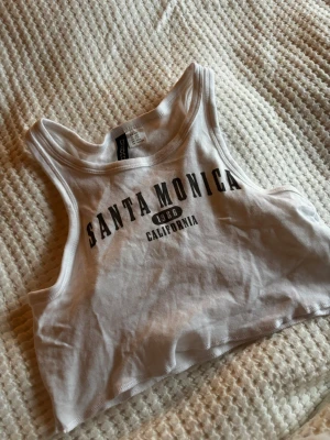 Vit croppad linnetopp Santa Monica - Säljer en vit croppad linnetopp från Divided med trycket 'Santa Monica 1986 California' i svart. Toppen är ärmlös och har en sportig, avslappnad stil med rundad halsringning. Perfekt för varma dagar och chill häng.