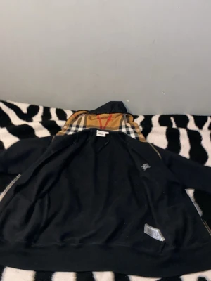 Svart hoodiejacka från Burberry - Svart hoodiejacka från Burberry med klassisk rutig huva i beige, svart, vit och röd. Jackan har dragkedja framtill och ribbade muddar. Perfekt för dig som vill ha en stilren look med en touch av lyx. Storlek “XS” men passar även 170 till 175 cm. Helt ny köpte för mindre än en månad. Jag kommer sälja den jätte billigt säkert under 1000kr så först som vill ha den för en pris kan jag sälja för 