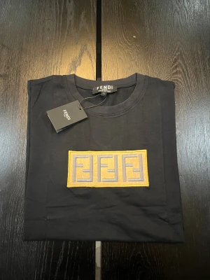 Svart Fendi t-shirt med broderad logga - Snygg svart t-shirt från Fendi i 100% bomull. På bröstet sitter en broderad patch med Fendis ikoniska logga i grått och gult. Klassisk rund hals och korta ärmar. Perfekt för dig som gillar exklusiva streetwear-vibbar och vill sticka ut med designerplagg.
