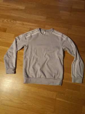 Ljusgrå Adidas sweatshirt med ränder - Säljer en ljusgrå Adidas sweatshirt med klassiska vita ränder längs ärmarna och Adidas-logga på axlarna. Tröjan har rund hals, ribbade muddar och är långärmad. Perfekt för en sportig och avslappnad stil.